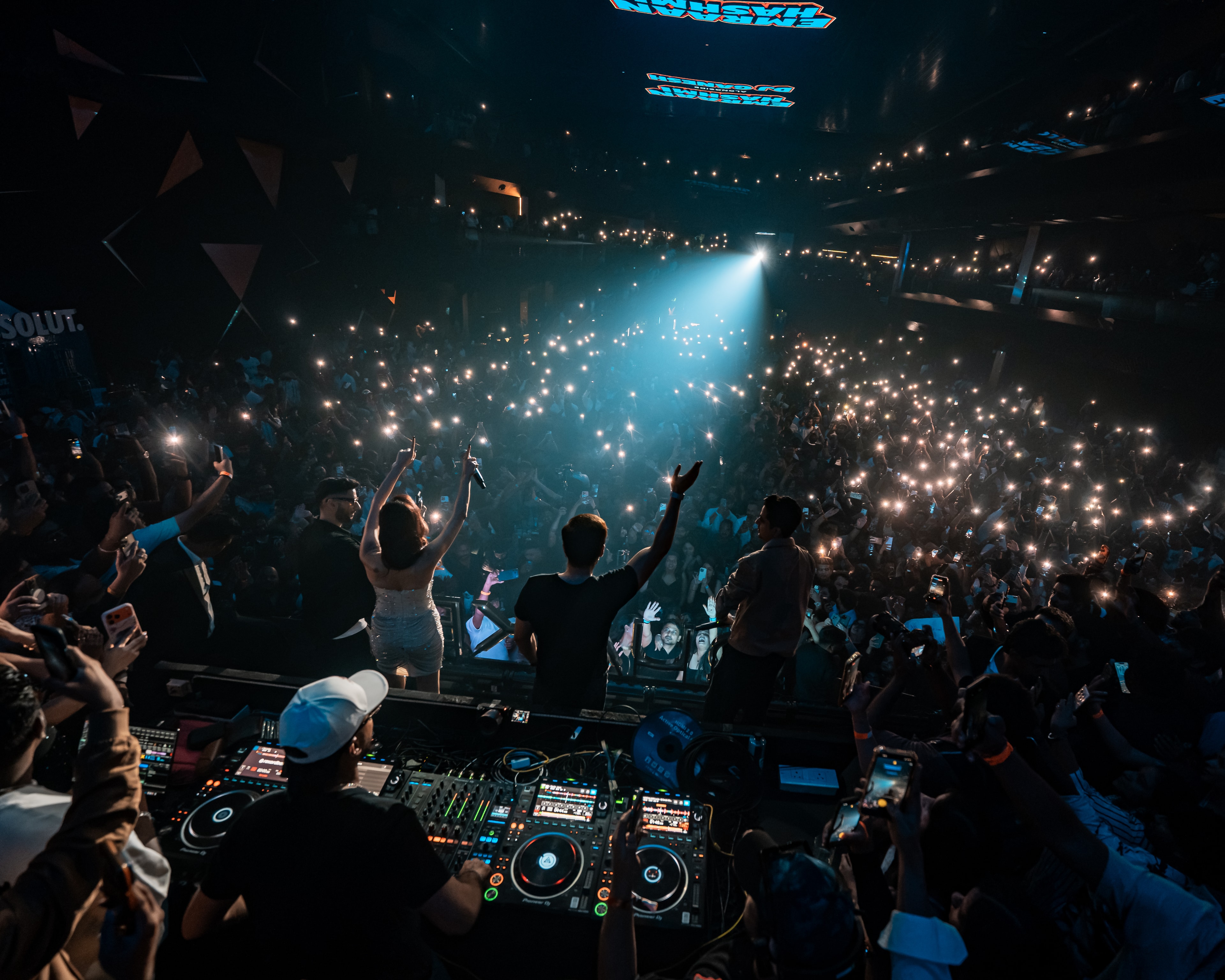 A packed arena lit by phone lights, seen from behind DJ Ganesh at the decks